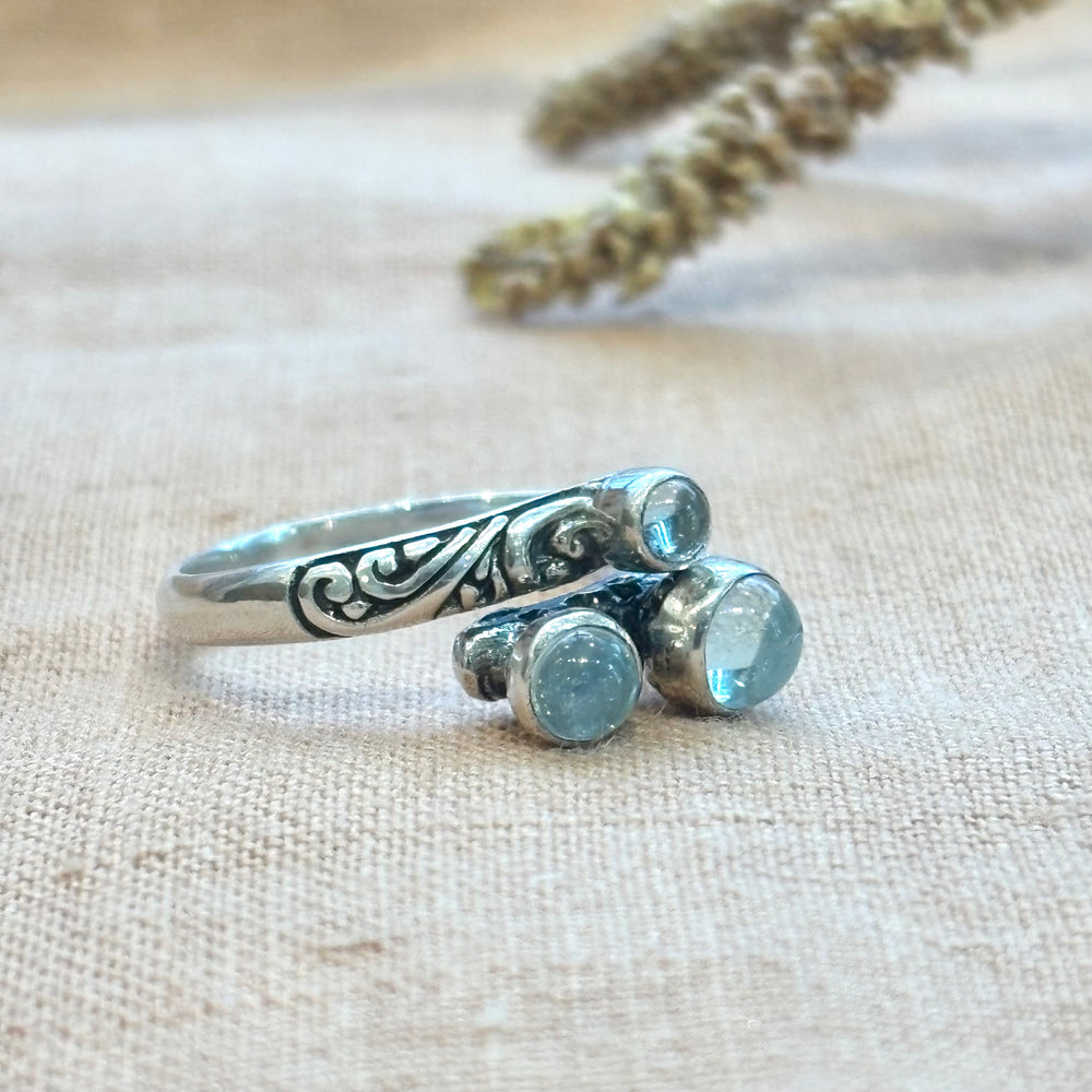 Three round aquamarine cabochons set in a hand-carved sterling silver scroll band ring, photographed on a natural linen background