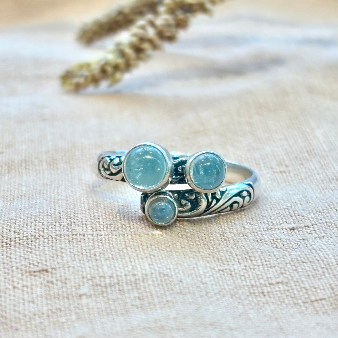 Three round aquamarine cabochons set in a hand-carved sterling silver scroll band ring, photographed on a natural linen background