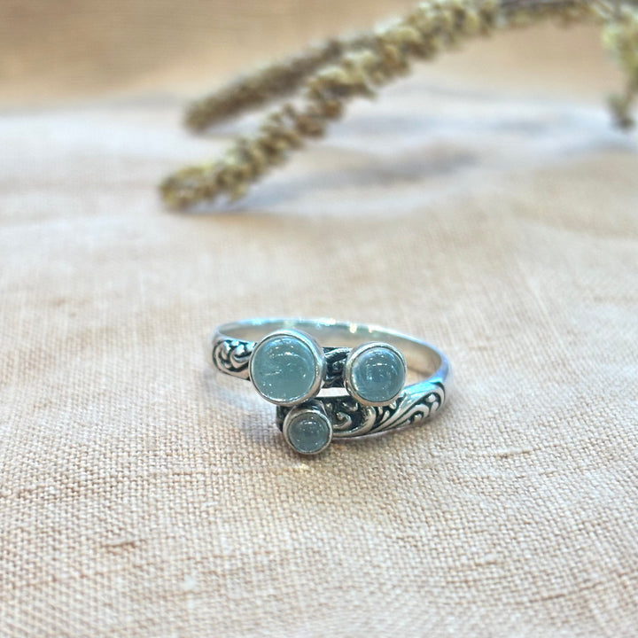 Three round aquamarine cabochons set in a hand-carved sterling silver scroll band ring, photographed on a natural linen background