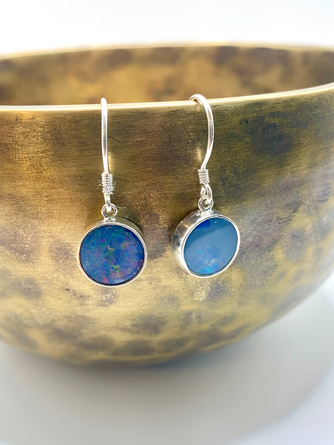 Pair of round blue opal drop earrings resting against a brass singing bowl, showing iridescent teal and violet stone tones