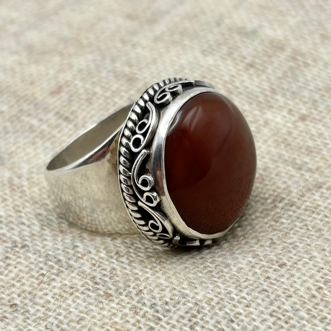 Large oval carnelian statement ring in sterling silver with hand-carved rope and scroll border detail, photographed at an angle on linen background