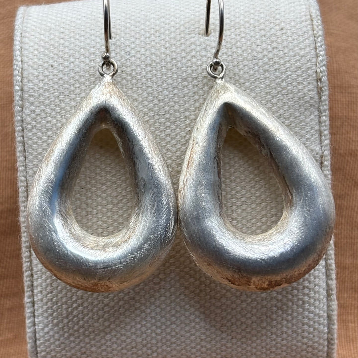 Close-up of large handmade sterling silver open teardrop earrings on a linen display stand, showing brushed texture and sculptural form