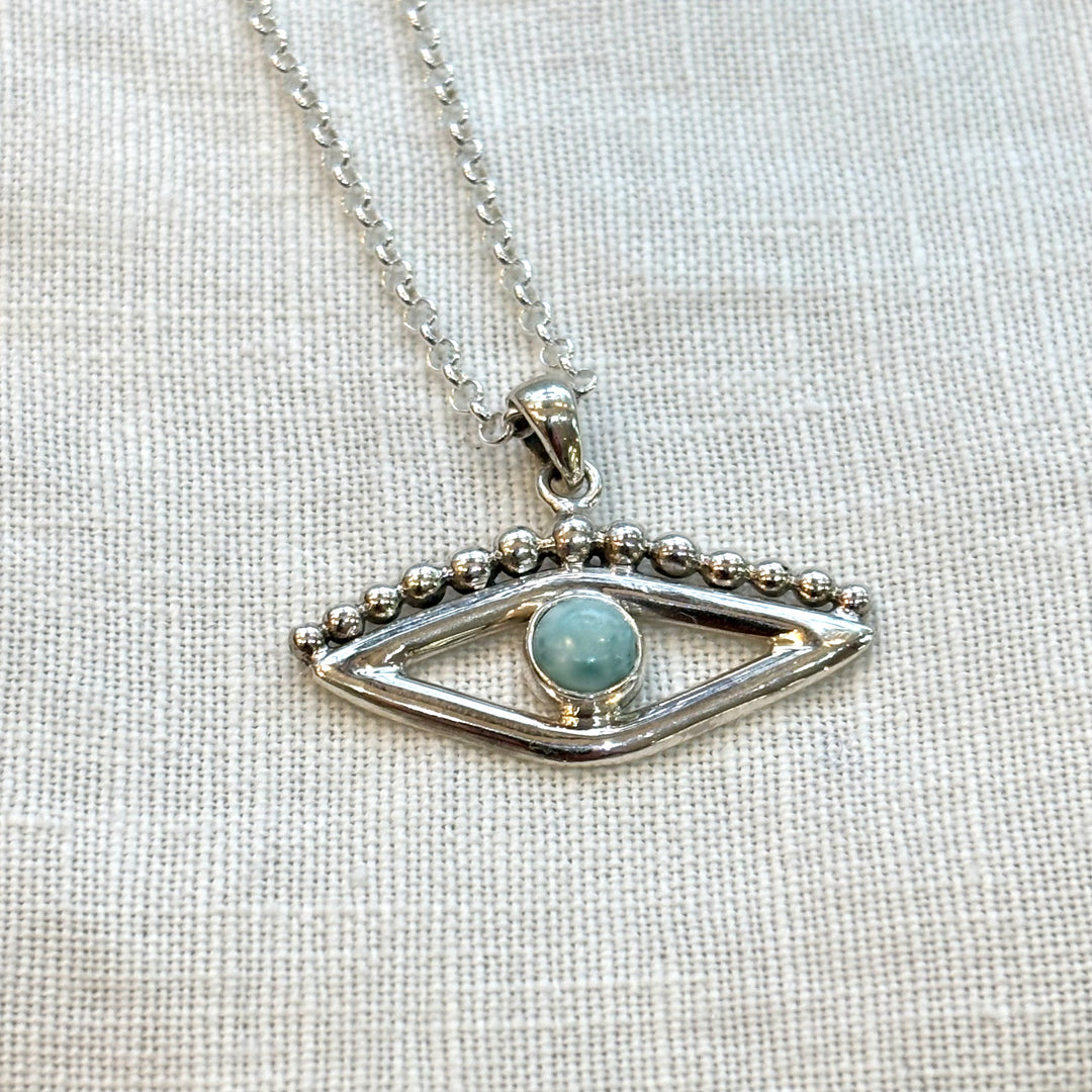 Close-up of a handmade sterling silver evil eye pendant set with a round blue larimar stone and silver bead detail, on a linen background