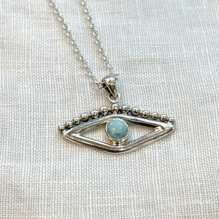 Close-up of a handmade sterling silver evil eye pendant set with a round blue larimar stone and silver bead detail, on a linen background