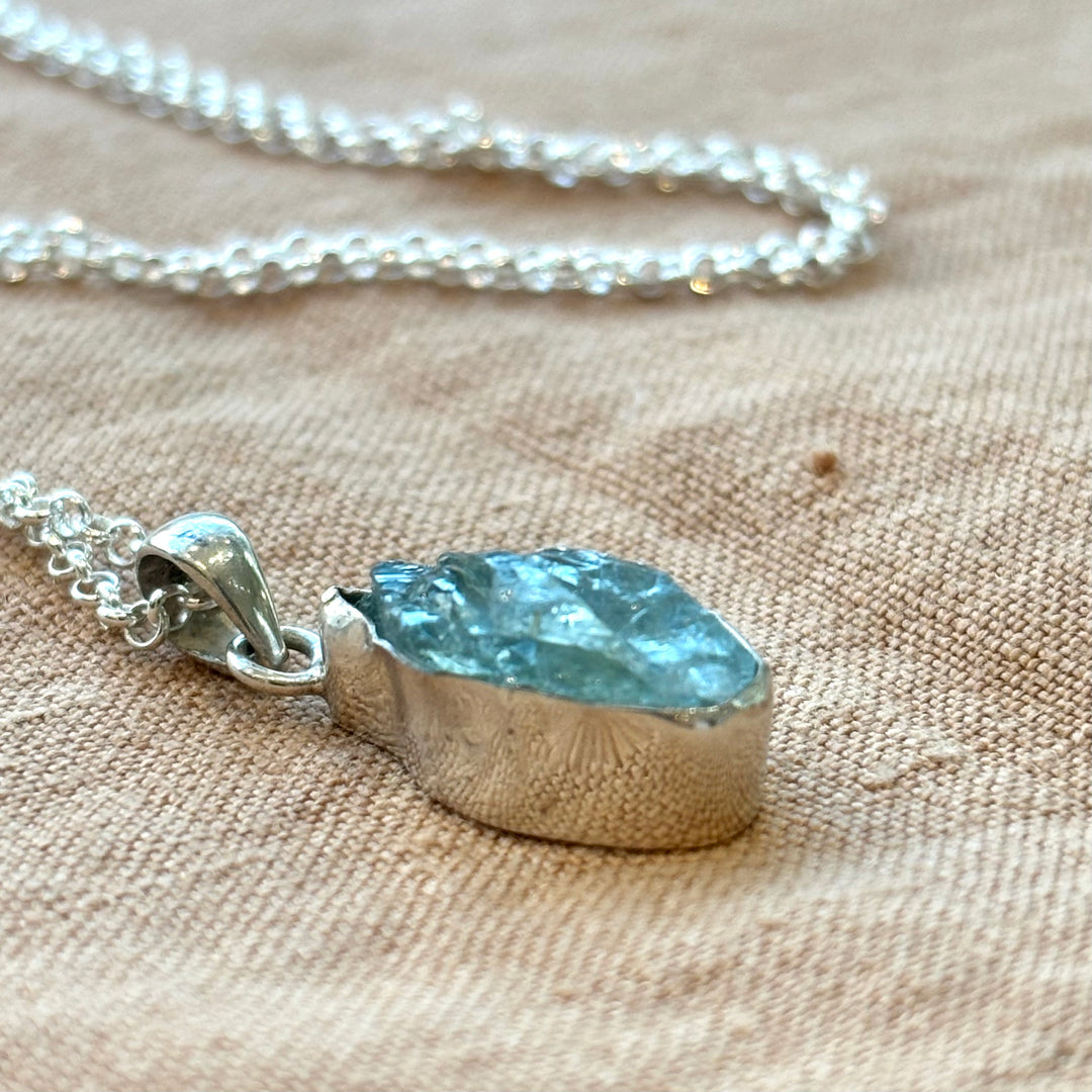 Close-up of a raw aquamarine crystal pendant set in a handmade sterling silver bezel on a linen background, showing natural blue-green stone texture and chain detail