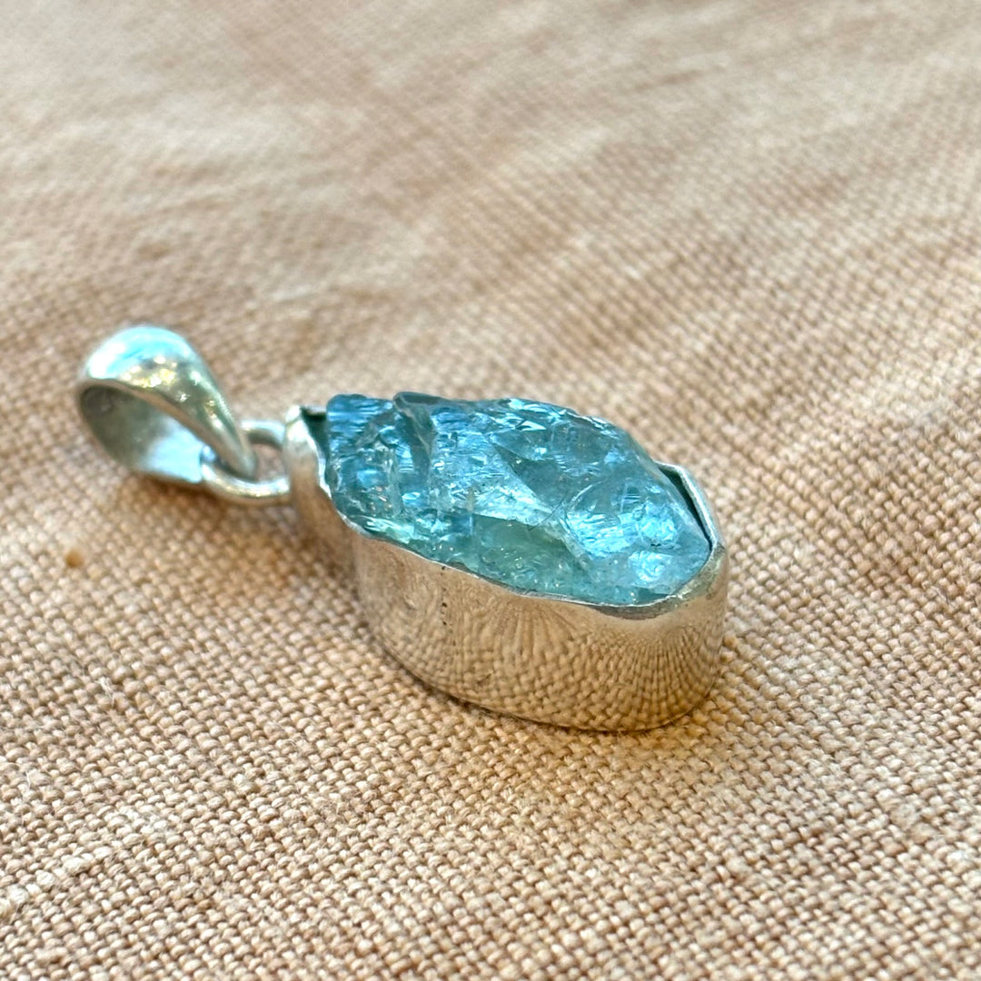 Angled view of a raw aquamarine crystal set in a handmade sterling silver pendant, showing the natural organic stone form and setting depth