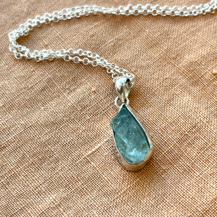 Top-down view of a raw aquamarine crystal sterling silver necklace showing the full pendant and 18 inch chain on a linen background